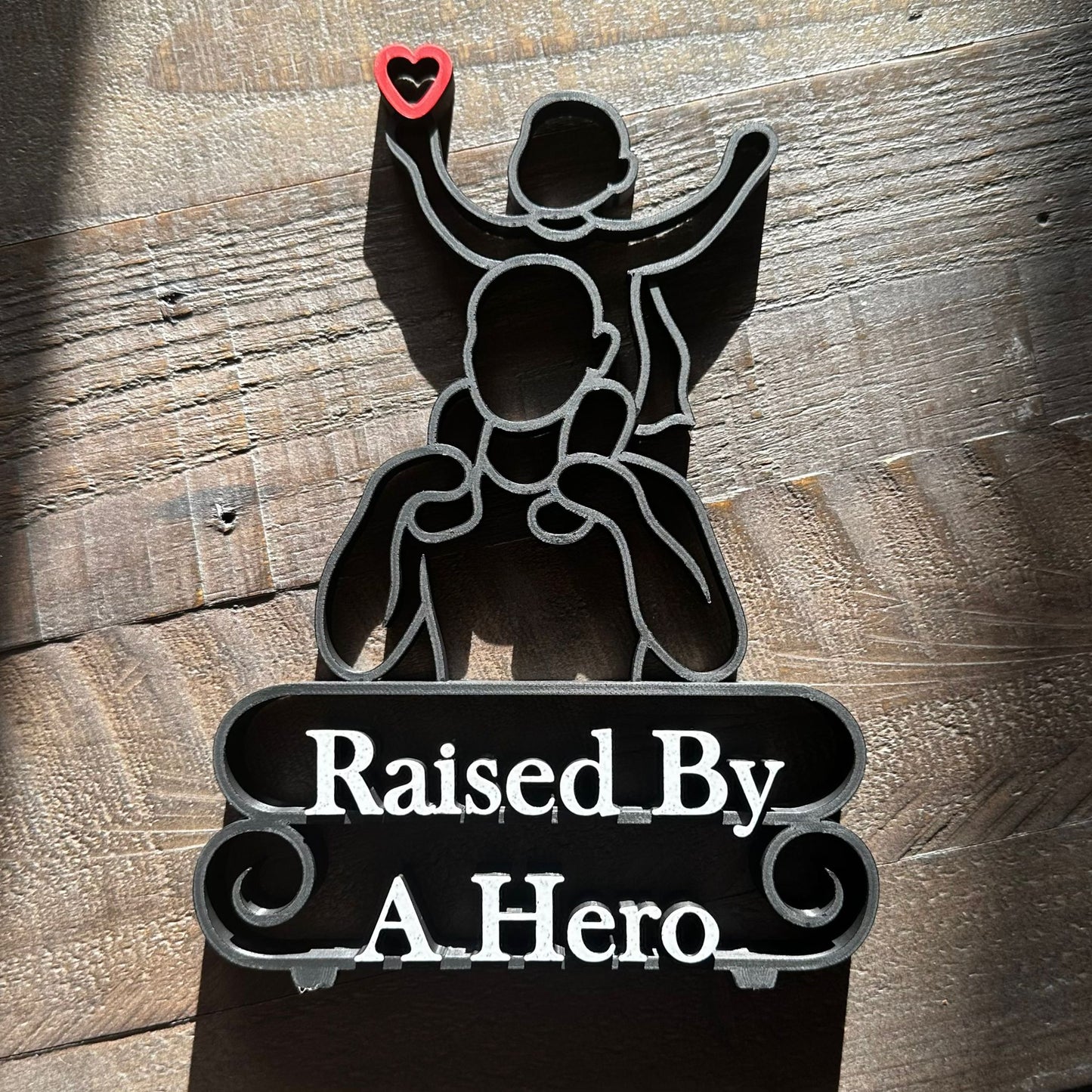 Modern line art sculpture with silhouette of a child on a father's shoulders and text 'Raised By A Hero' below. Black, with red heart and marble text. Lying down on a wooden background.