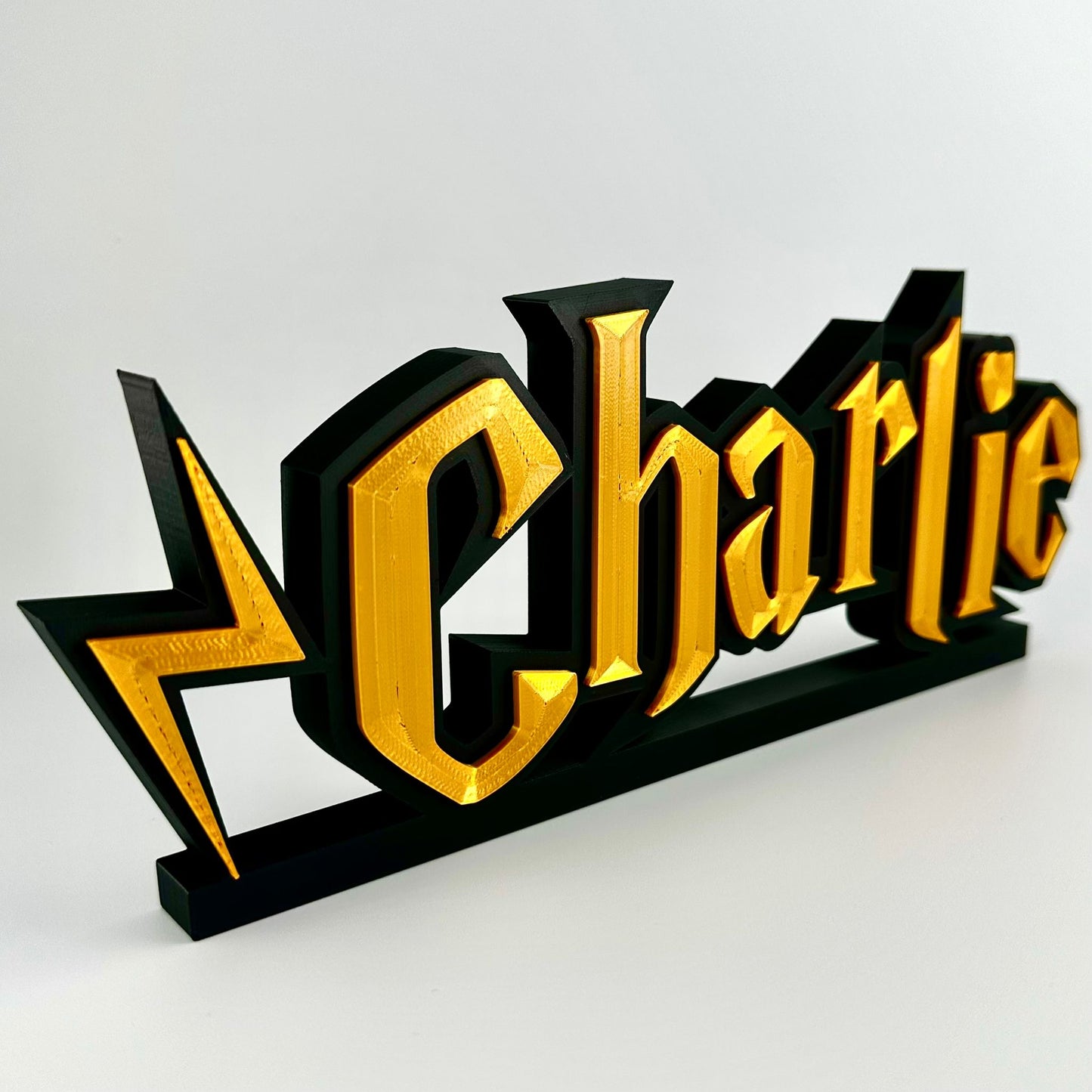 A custom made nameplate in a wizard style font, showing the name Charlie. Gold 3d text on matte black background, freestands upright on a black base.