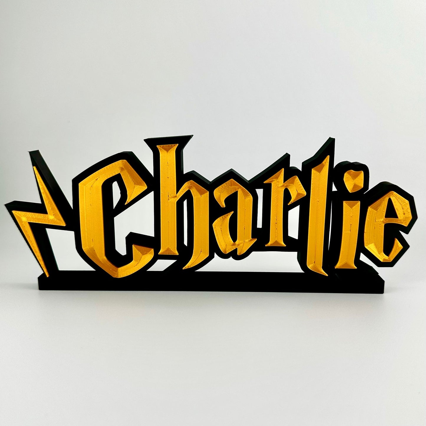 A custom made nameplate in a wizard style font, showing the name Charlie. Gold 3d text on matte black background, freestands upright on a black base.