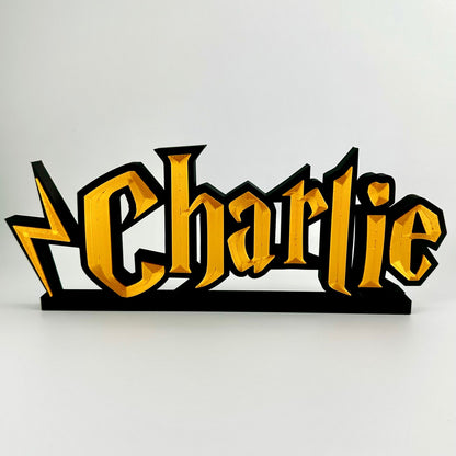 A custom made nameplate in a wizard style font, showing the name Charlie. Gold 3d text on matte black background, freestands upright on a black base.
