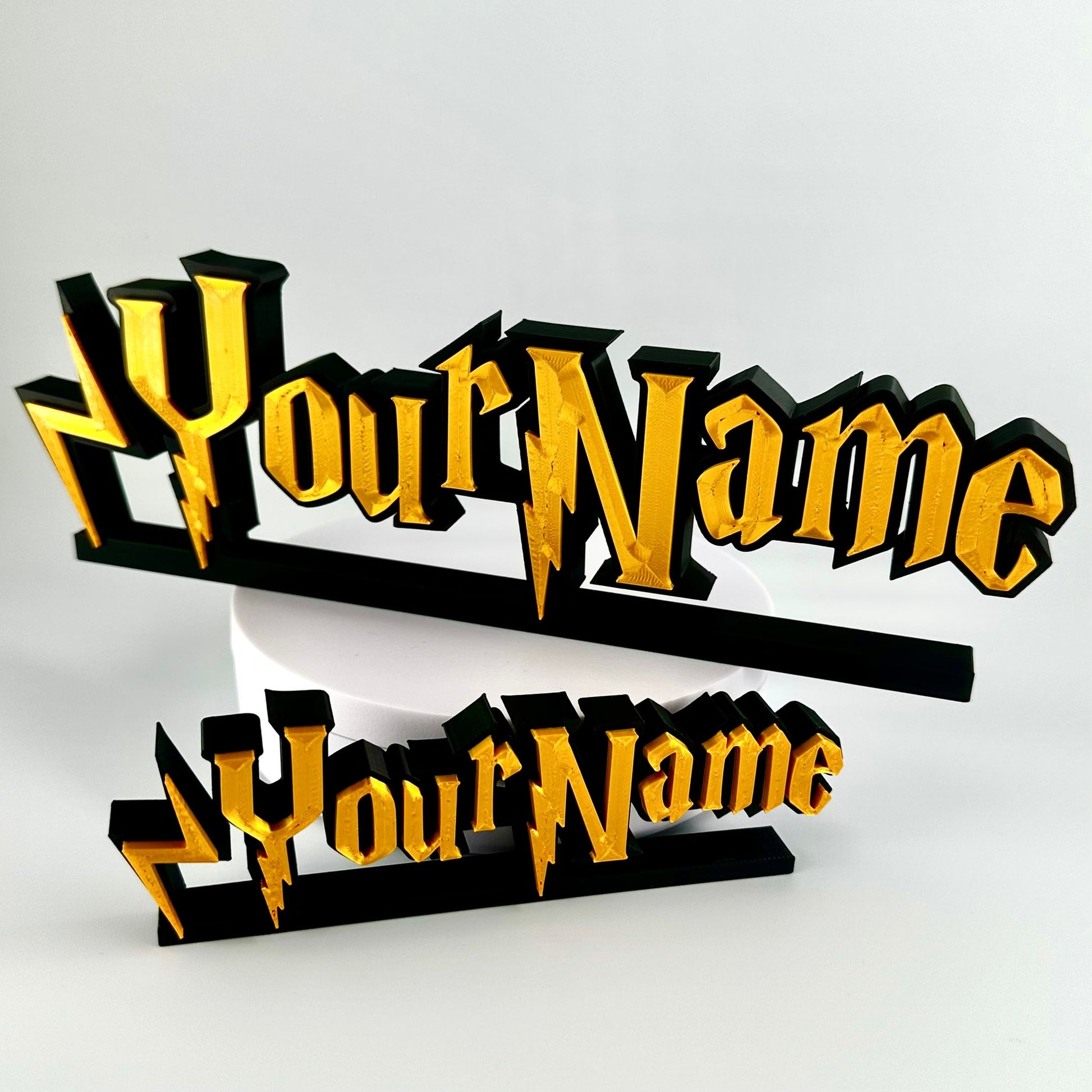 Custom made nameplates in a wizard style font. Two variations are shown, one 15cm wide the other 25cm wide.  Gold 3d text on matte black background, freestands upright on a black base.