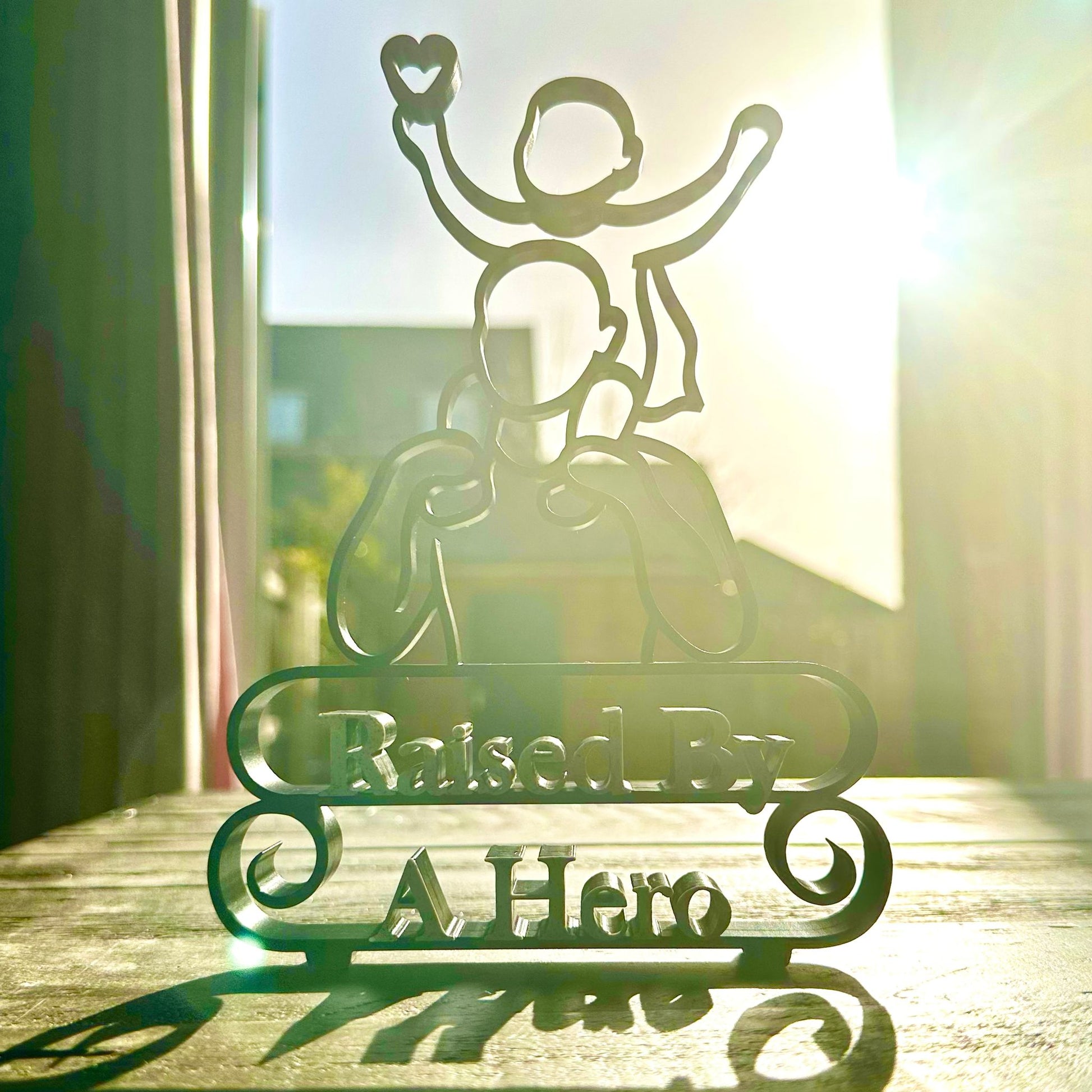 Modern line art sculpture with silhouette of a child on a father's shoulders and text 'Raised By A Hero' below. Black, with red heart and marble text. Freestanding on a table, backlit by the sun.
