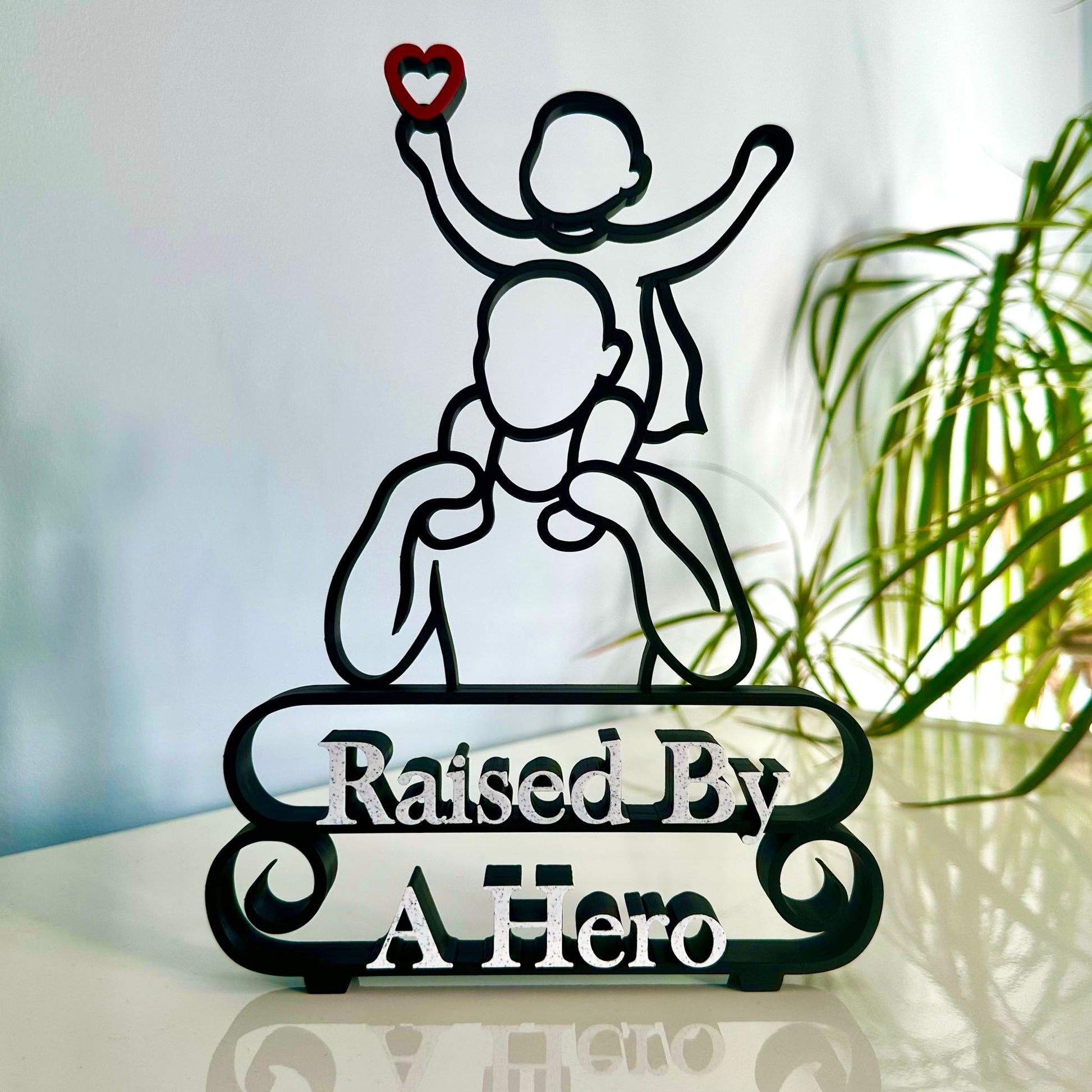 Modern line art sculpture with silhouette of a child on a father's shoulders and text 'Raised By A Hero' below. Black, with red heart and marble text.