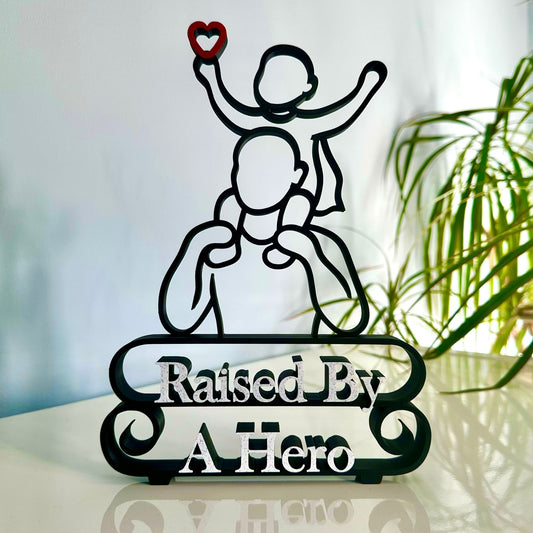 Modern line art sculpture with silhouette of a child on a father's shoulders and text 'Raised By A Hero' below. Black, with red heart and marble text.