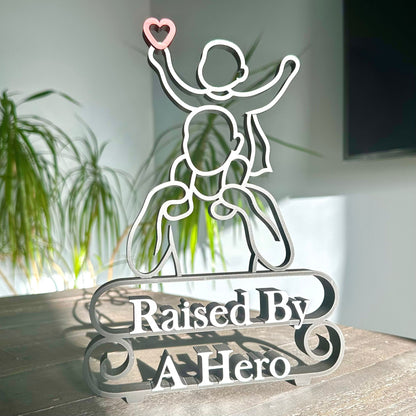 Modern line art sculpture with silhouette of a child on a father's shoulders and text 'Raised By A Hero' below. Black, with red heart and marble text.