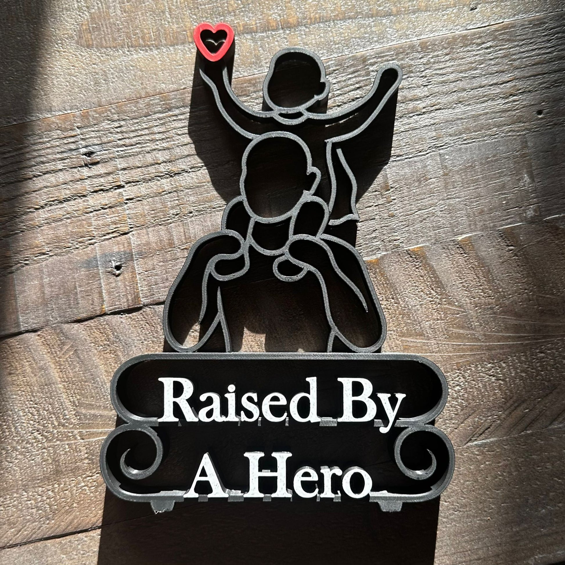 Modern line art sculpture with silhouette of a child on a father's shoulders and text 'Raised By A Hero' below. Black, with red heart and marble text. Lying down on a wooden background.