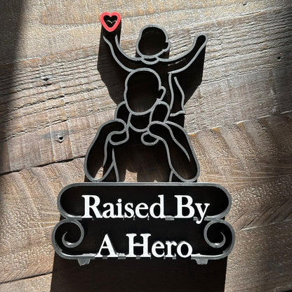 Modern line art sculpture with silhouette of a child on a father's shoulders and text 'Raised By A Hero' below. Black, with red heart and marble text. Lying down on a wooden background.