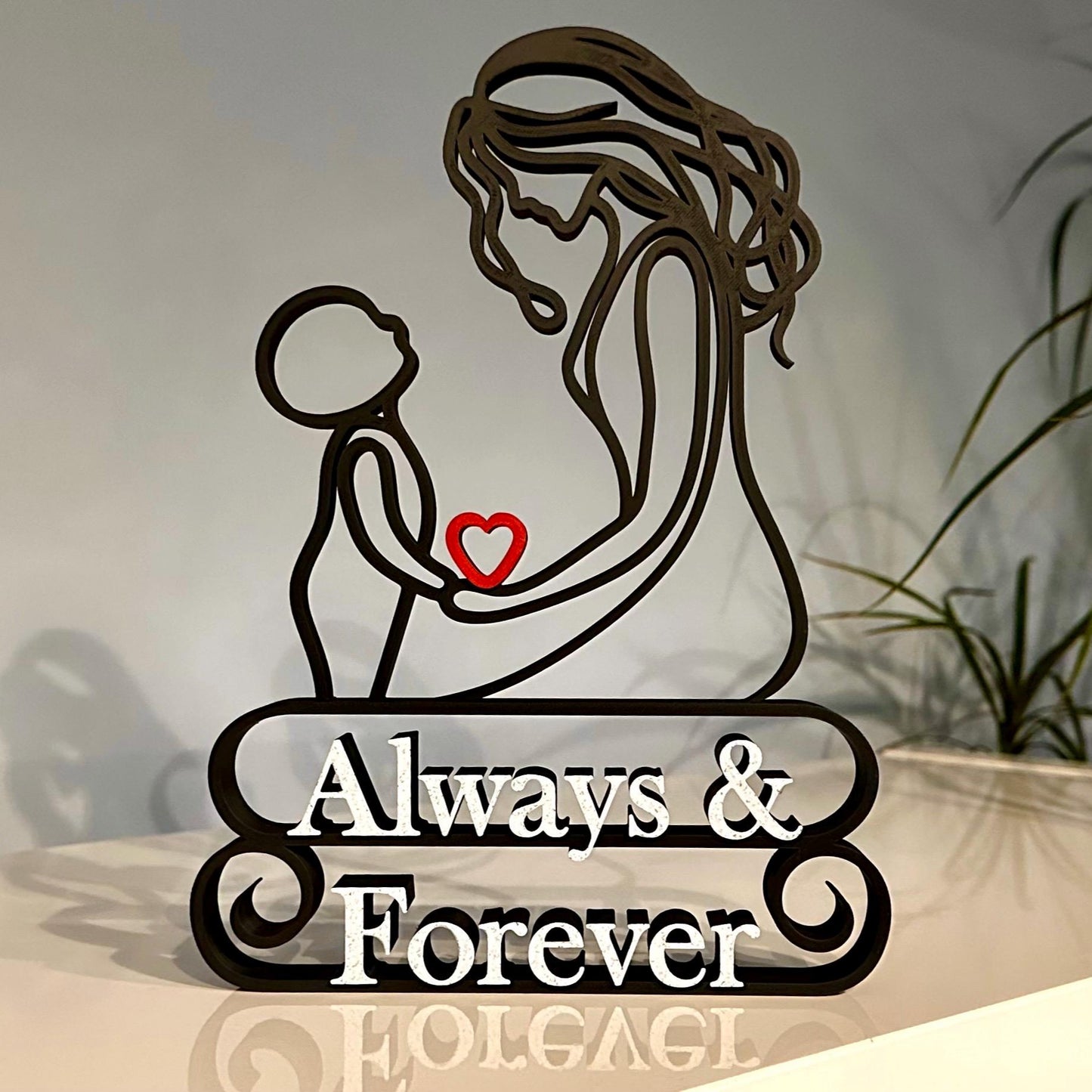 A line art sculpture of a mother and child with a red heart and the text 'Always & Forever' on the base, made from matte black or grey PLA with a marble effect finish.