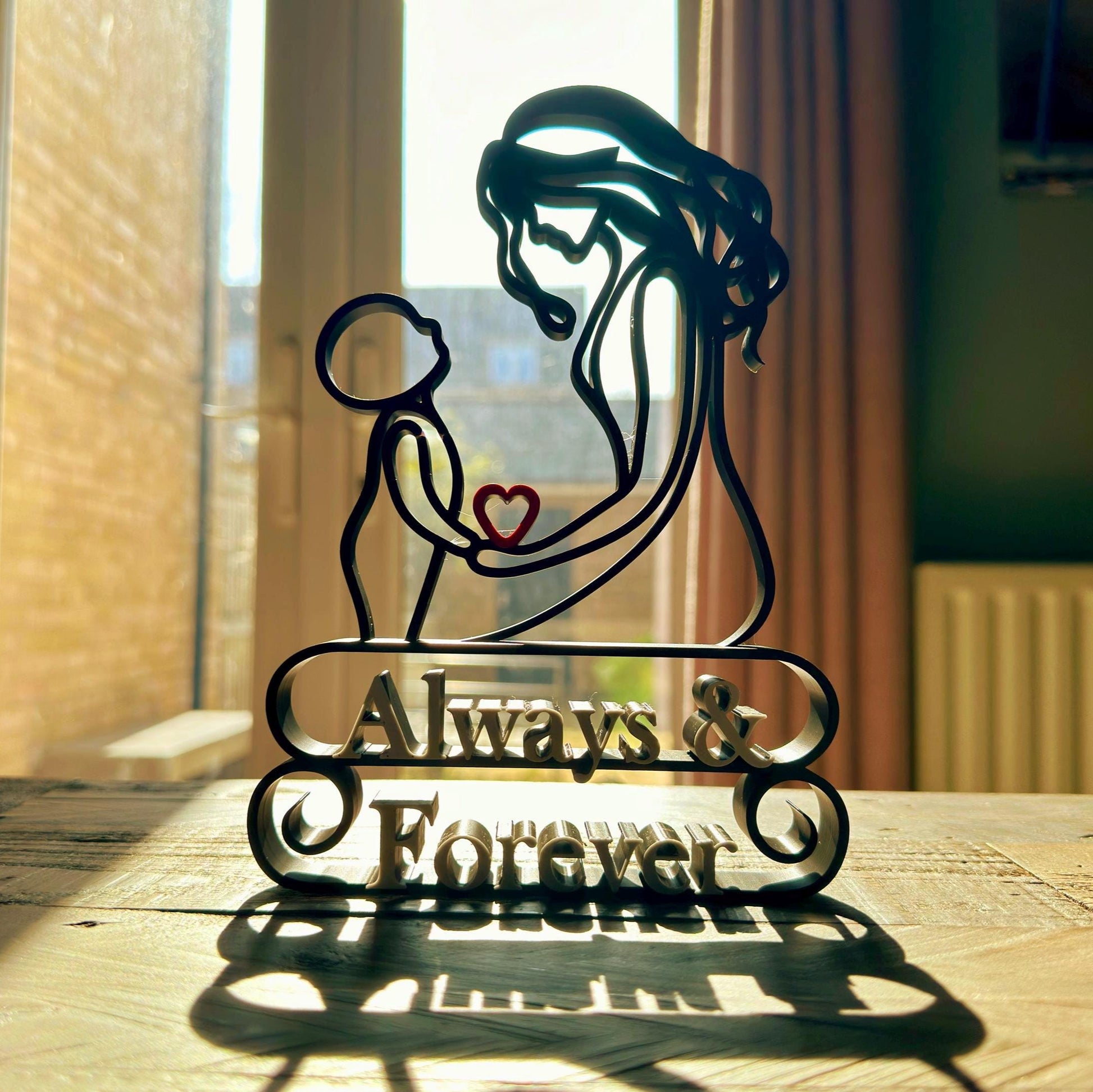A line art sculpture of a mother and child, with a heart shape between them, placed on a freestanding base which has the inscription 'Always & Forever'. The sculpture is on a table with natural light coming from a window in the background.