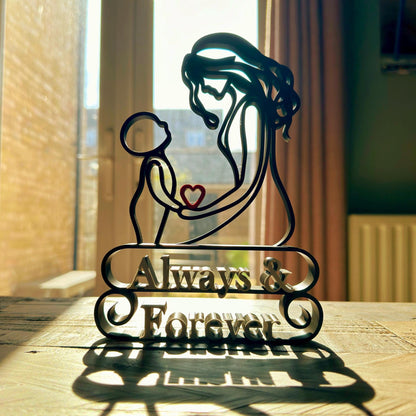 A line art sculpture of a mother and child, with a heart shape between them, placed on a freestanding base which has the inscription 'Always & Forever'. The sculpture is on a table with natural light coming from a window in the background.